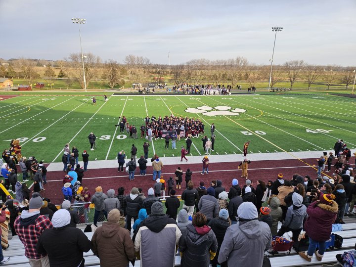A crowd gathers in the stands at Big Cat Stadium, with fans bundled up for the cool weather. On the field, players in maroon and black uniforms huddle near the 30-yard line, surrounded by team staff and fans. The large paw logo is visible at midfield, with trees and open landscape in the background under a cloudy sky.